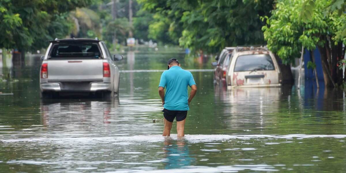 A man in a teal shirt and black cap wades through deep floodwaters on a street with submerged silver pickup and rusty white car.