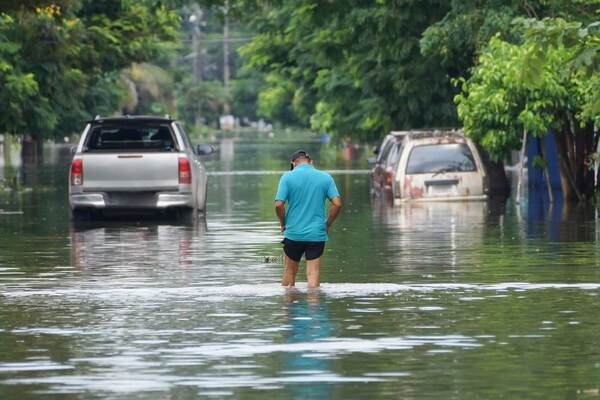A man in a teal shirt and black cap wades through deep floodwaters on a street with submerged silver pickup and rusty white car.